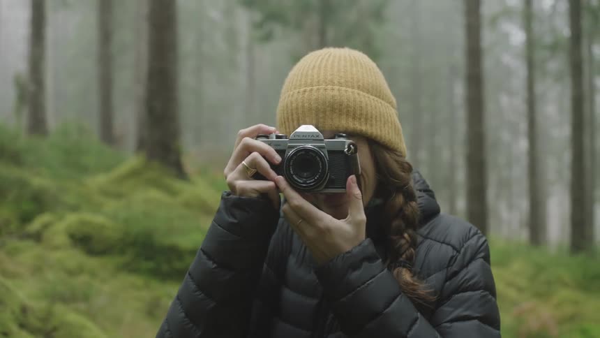 A woman takes a picture with her camera while she is laughing standing in a forest during the day
