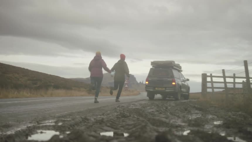 A man and a woman run along a countryside road towards a vehicle with flashing emergency lights on the side of the road before climbing in on a cloudy day
