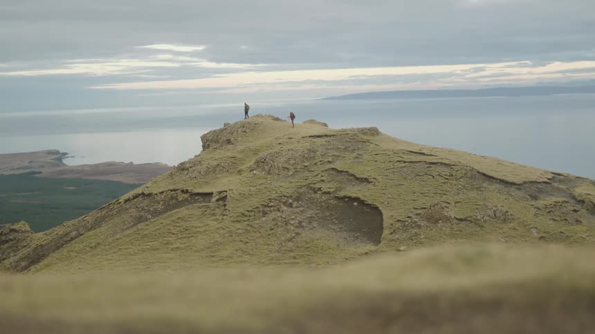 A hiker standing with another hiker on a plateau high in the mountains chases and dives after a dropped sheet of paper being blown towards the plateaus edge on a cloudy evening