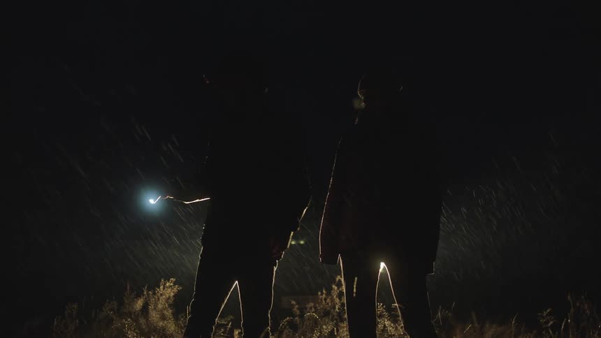 A man holding a flashlight is walking with a woman through tall grass on a rainy night with a car behind them