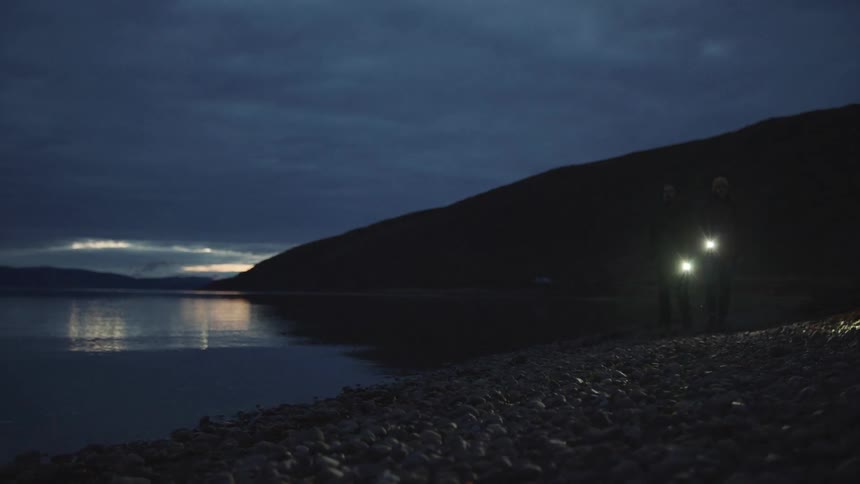 A man and woman walk along the shore at night while carrying flash lights