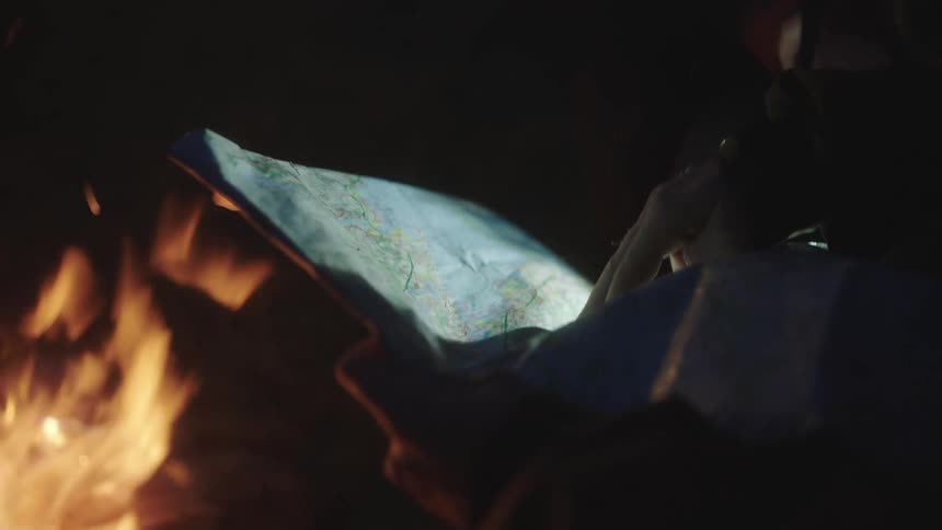 A man and a woman look at and point at a map with a campfire nearby