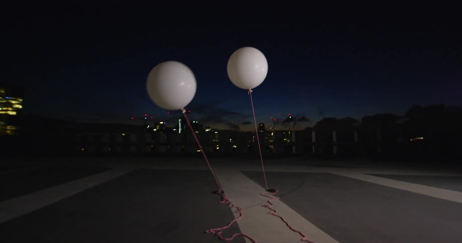 Two circular white balloons float on pink ropes that are taped to the ground across from downtown