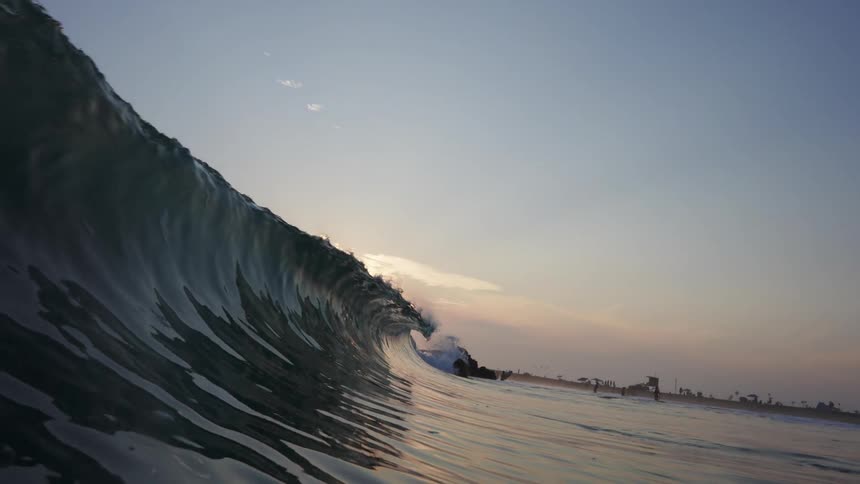 Ocean wave crashes with people in the distance on beach with umbrellas at sunset