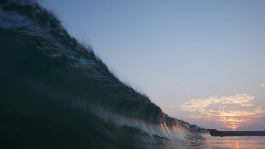 A wave crashes in an ocean as the sun sets in the distance