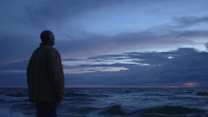 Middle aged man with a coat and pants stands and watches the sea at dusk