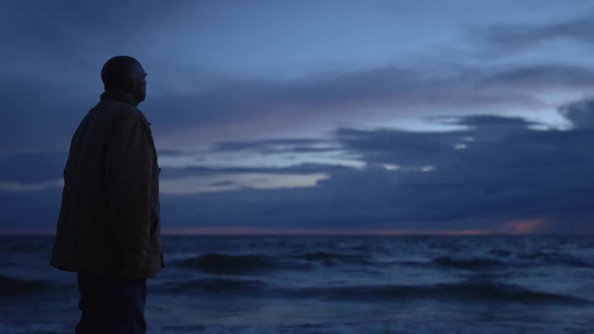A bald man stands at the shore of the ocean as day turns to night