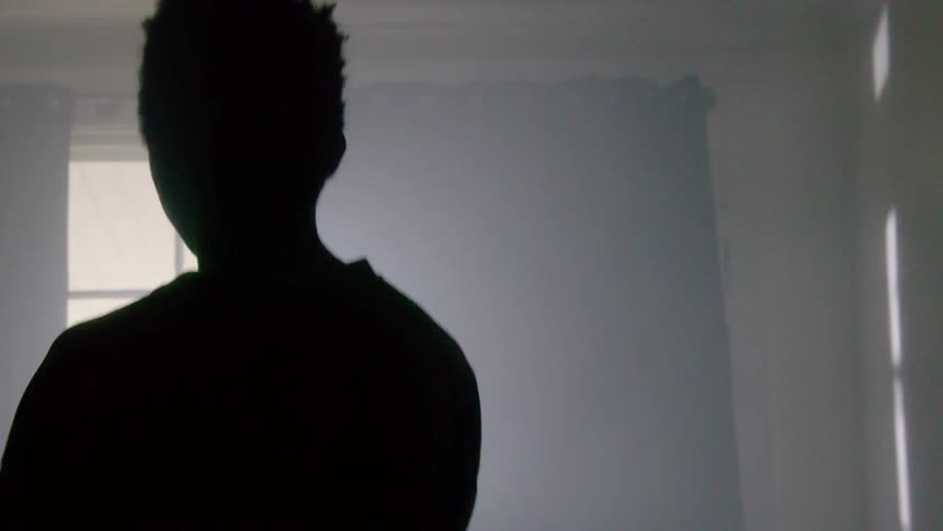 A young man jumps and dances around a room in front of an open window