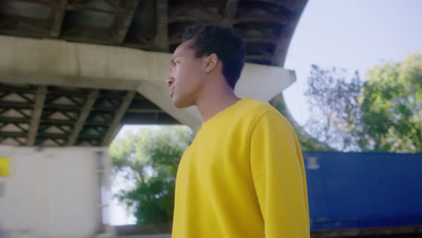 Young black man wearing a yellow sweater takes a stroll down the street under an overpass with a train in the background