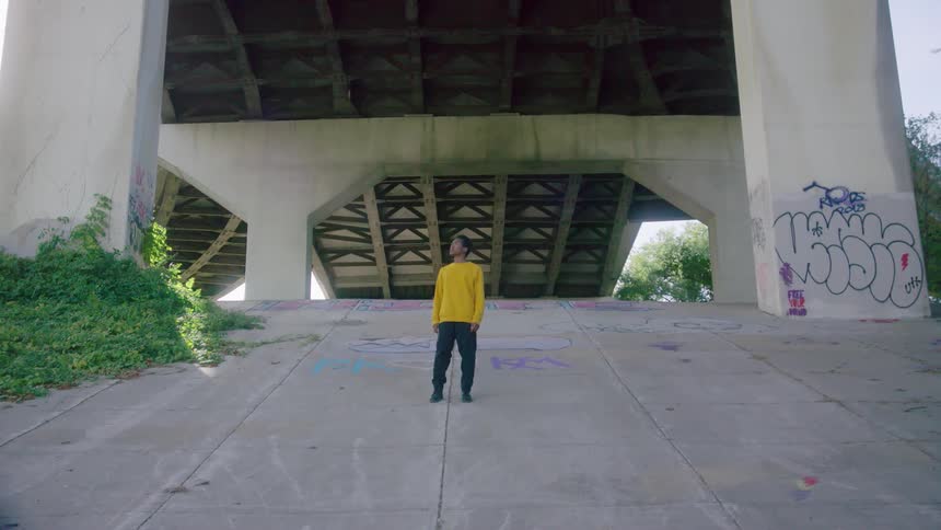 Boy standing on on a slope staring off to the side at daytime under a bridge with graffiti