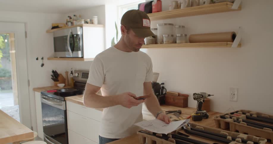 A young white man is standing in his kitchen trying to figure something out while looking at some instructions and his smart phone