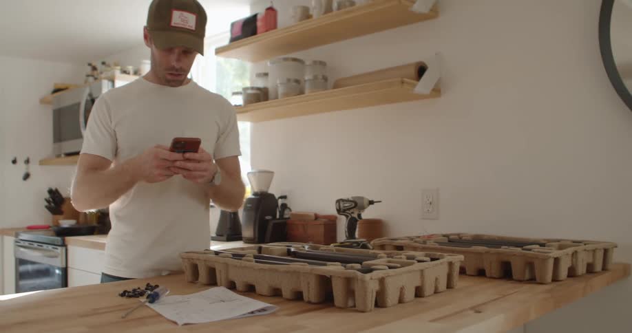 A man looks at metal items inside cardboard packaging in his kitchen and does a search on his smartphone