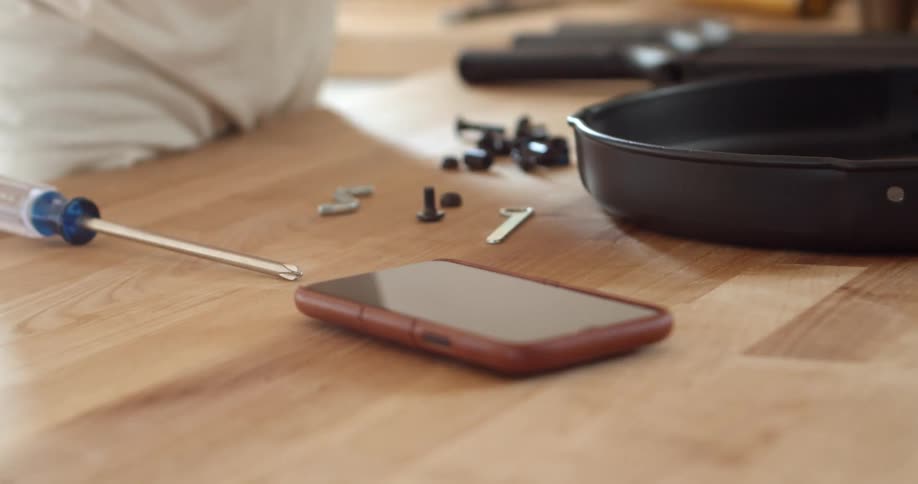 Cellphone with a red case on top of a counter in a home next to a man assembling screws and bolt pieces