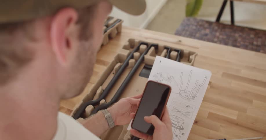 A man stands indoors holding a phone and looking at a piece of paper with a diagram