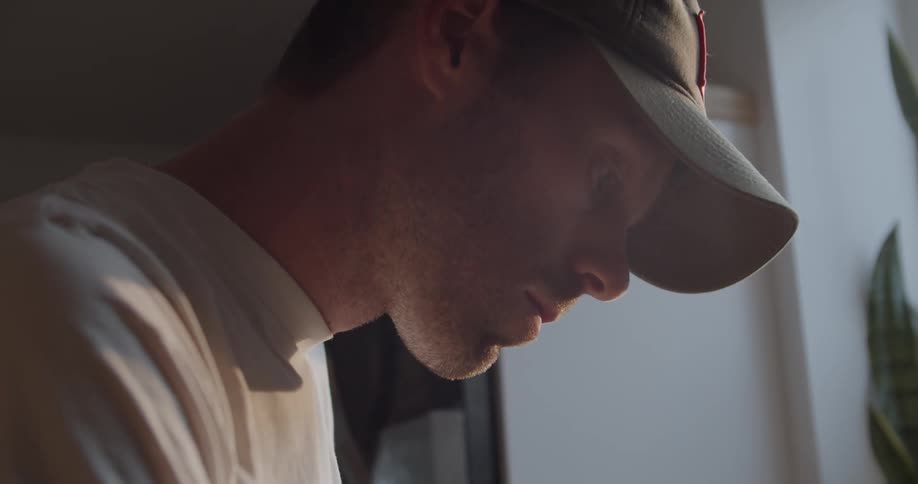 A man stands inside next to a window wearing a baseball hat and t shirt