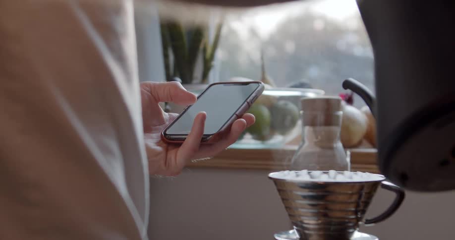 Man uses a smartphone while pouring water into a cup in the kitchen