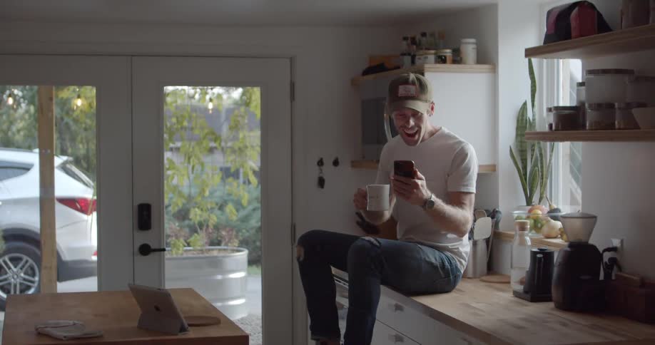 Man sitting on a countertop in a kitchen holding a coffee mug and a smart phone