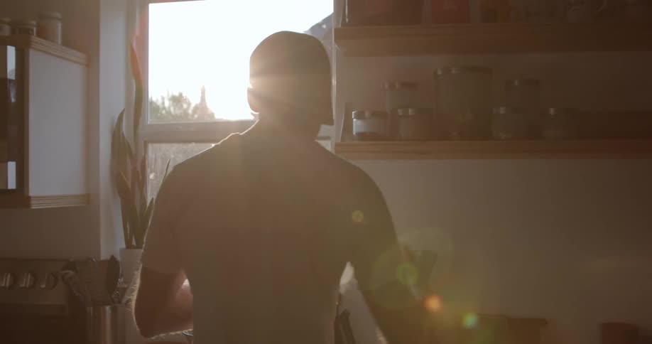 Man standing in his kitchen in front of a coffee machine at day time reaching over to a shelf to grab a coffee filter