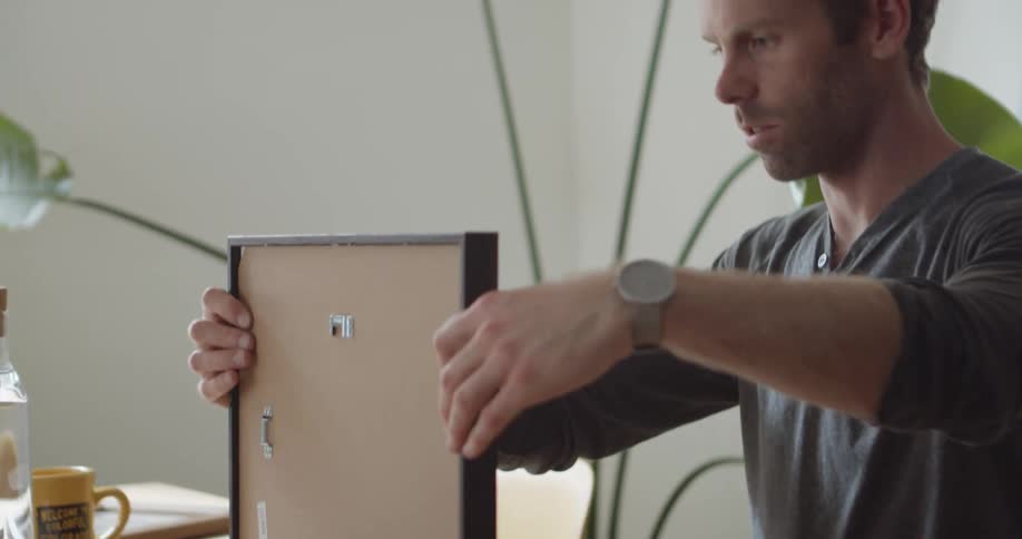 A man sitting at a desk turns a picture frame right side up to check the framing