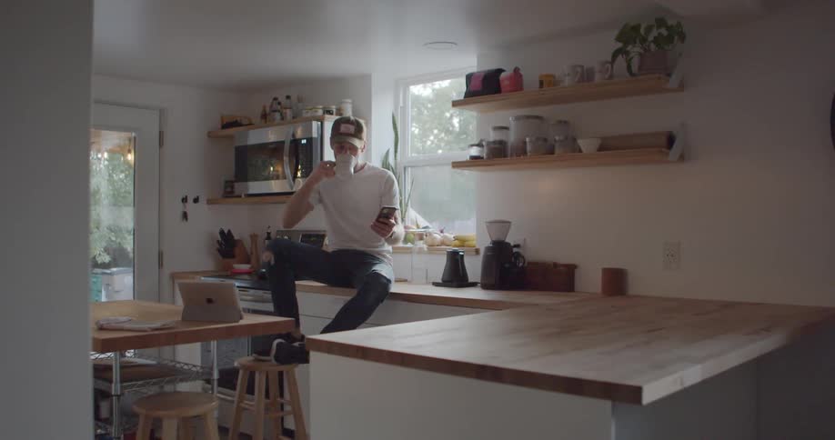 Man with a white shirt and a brown cap sitting on the kitchen counter uses a smartphone while drinking a cup of coffee in the kitchen