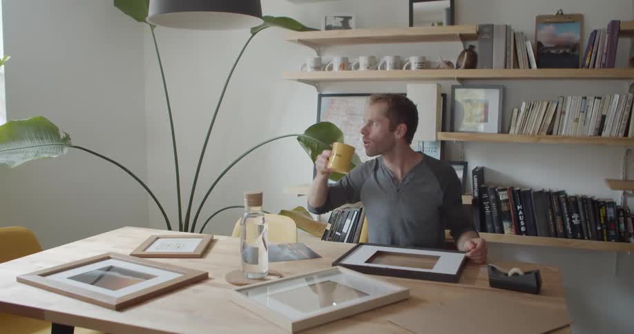 Man sitting at a dining room table at day time drinking from a yellow coffee mug and looking out a window