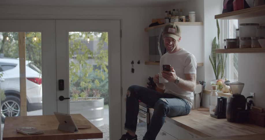 A male caucasian sitting in a kitchen on the counter using a cell phone