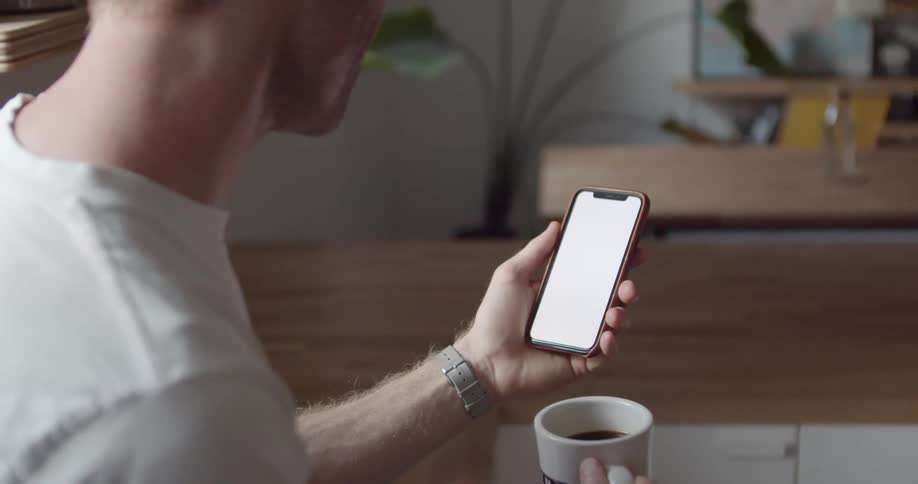 Man with a brown cap and a white shirt uses a smartphone and holds a up in the kitchen