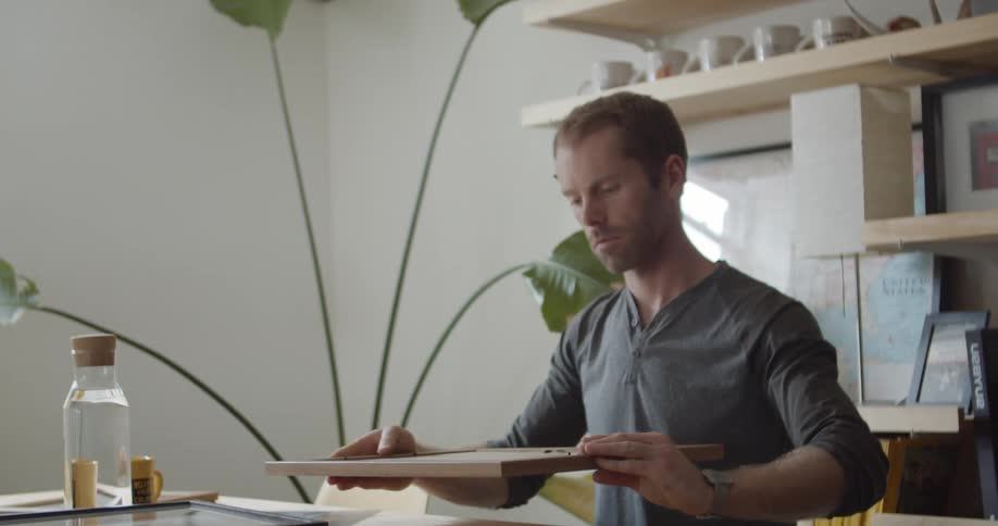 A turns a picture frame around and sits down at a table then starts to undo the back of the frame