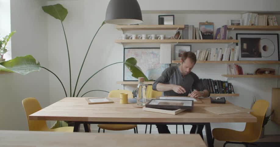A man sits at his kitchen table and tapes a paper print to frame