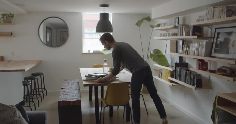 A man carries a stack of picture frames and places them on a table in a home dining room before taking a seat