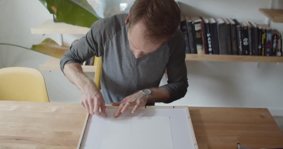 Man sitting at a table in his house using tape to tape a piece of paper in place on a frame