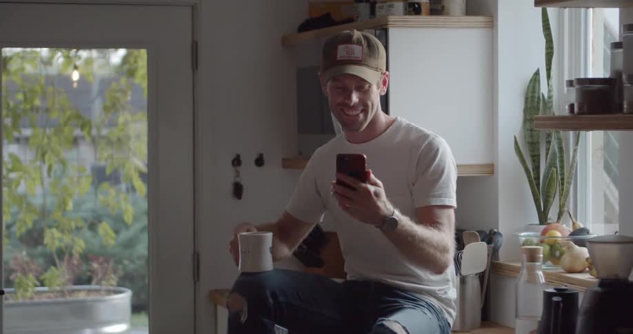 A man smiles as he sits on his kitchen counter and talks on his smart phone