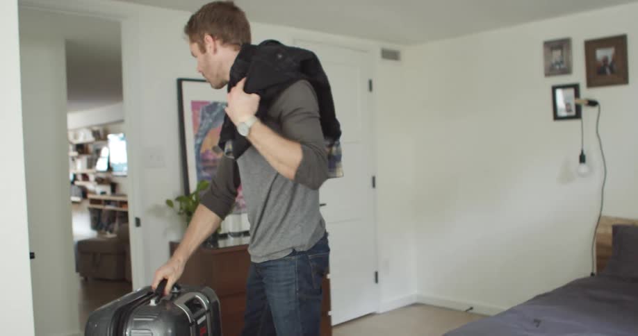 A man tosses clothing over his shoulder and then rolls his suitcase into an adjacent room
