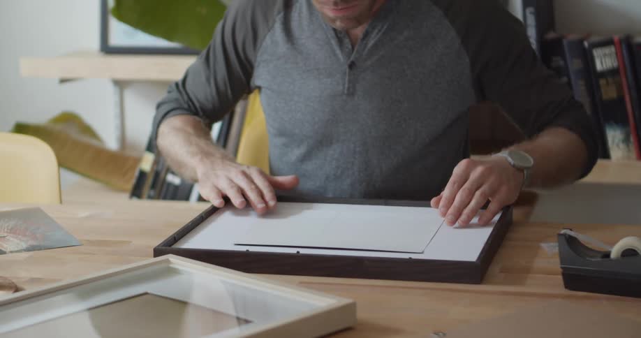 A man sits at a table placing a picture into a frame for hanging