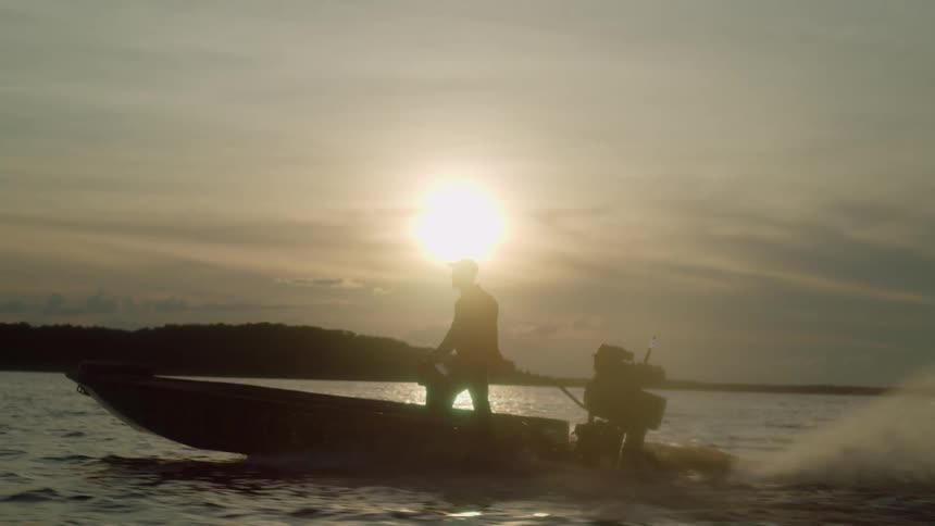 Man stands in boat piloting it across water as sun hovers in cloudy sky over distant shore