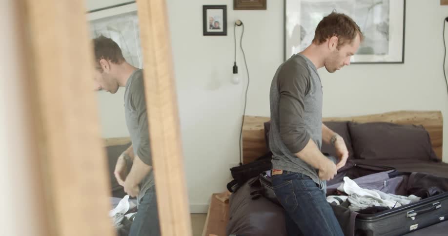 A man rolls clothing before packing it into an open suitcase on a bed