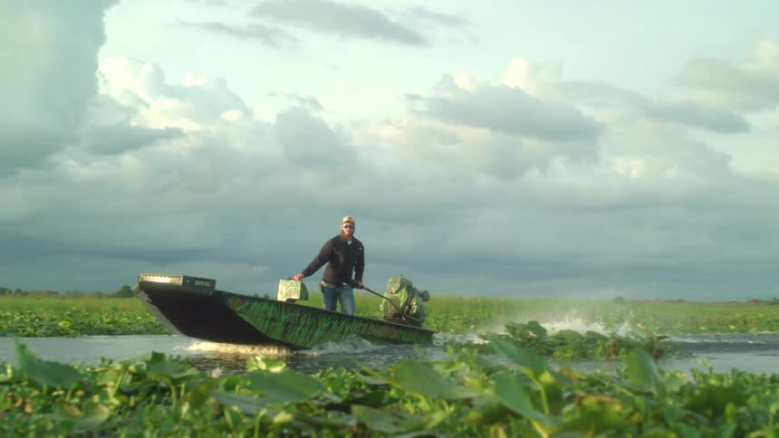 A person standing on a boat operating the motor while traveling on a river
