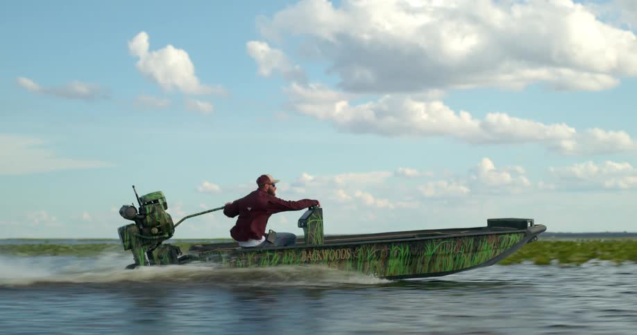 A man rides a motorboat with grass painted on the hull across a marsh on a sunny day