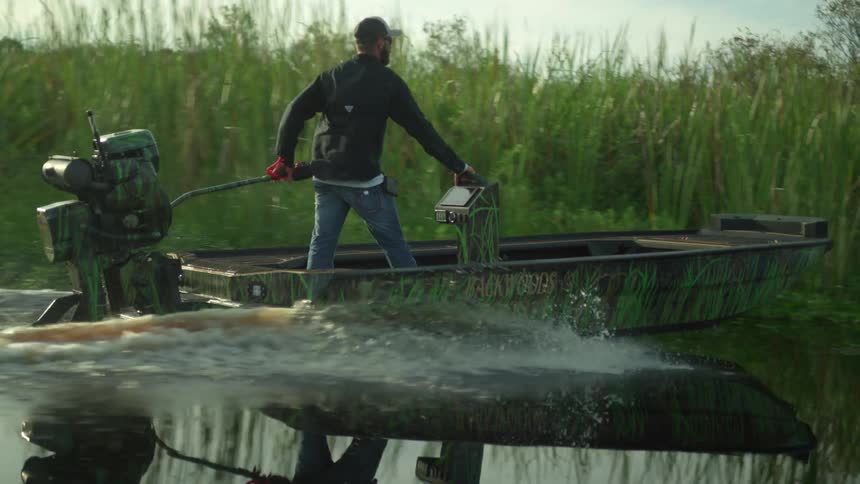 A man stands on a motor boat traveling through marsh water near tall grass on shore
