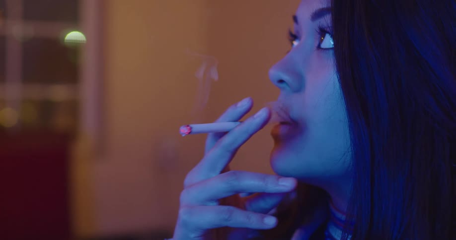 Woman with dark wavy hair smokes a cigarette in a room