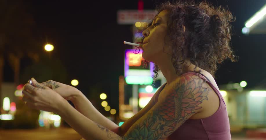 A woman stands outside on the street lighting a cigarette