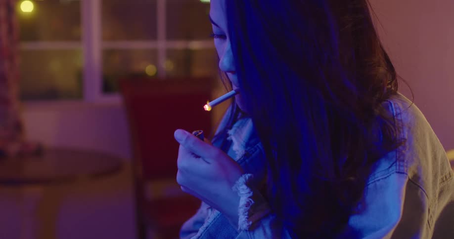 Woman standing in room under rose tinted light smokes a freshly lit cigarette