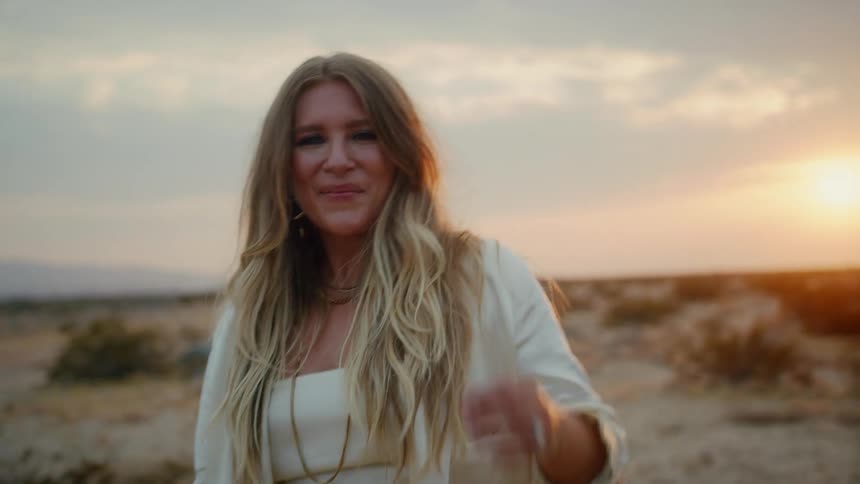 A young woman with long blonde hair dances in a desert as the sun sets