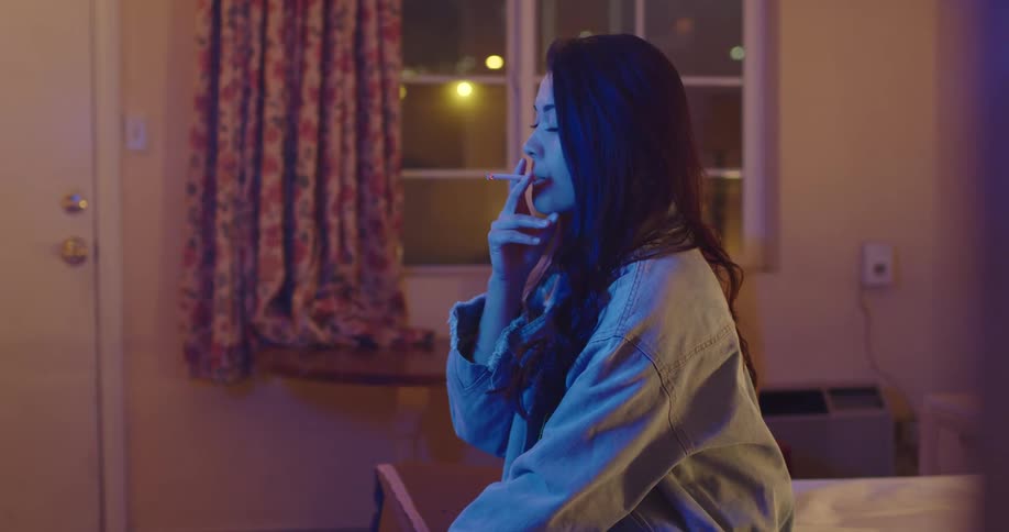 Woman sitting at the edge of a bed in a motel room at nighttime smoking a cigarette staring at a tv