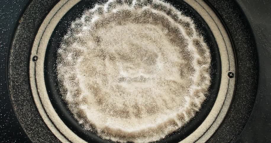 Sand in center of container vibrates shaking particles onto surroundings