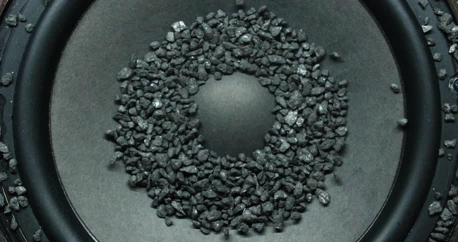 Small black rocks are moving off a speaker due to vibrations on the surface