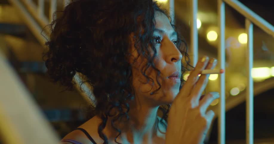 Tattooed woman with curly hair smokes a cigarette sitting on the stairs outdoors