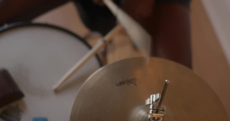 A man sitting on a stool before a drum set strikes the cymbal with a wooden drumstick