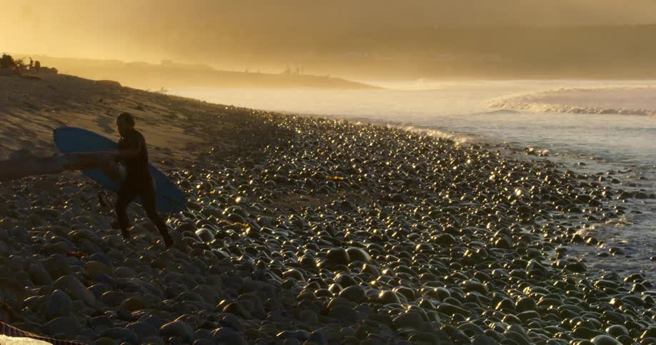 A surfer walking up a rocky shoreline at sunrise