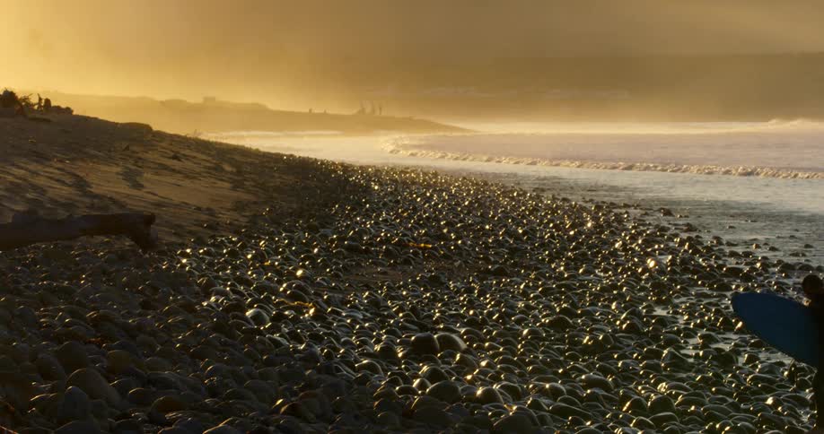 A surfer walking up a rocky shoreline on the coast at sunrise